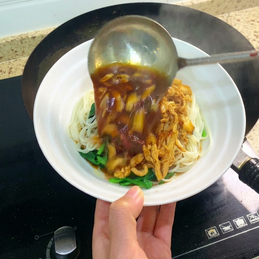 Une grande louche en métal versant une sauce sombre et savoureuse au poulet effiloché et aux champignons sur un bol blanc de nouilles.