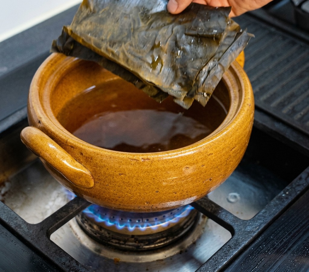 Une main déposant des feuilles de kombu séchées, vert foncé et rectangulaires, dans une marmite en argile brune placée sur la gazinière.