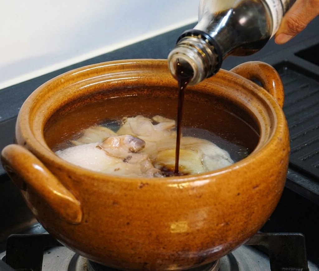 Une main versant un assaisonnement liquide sombre depuis une bouteille en verre dans la marmite contenant le poulet et leau.