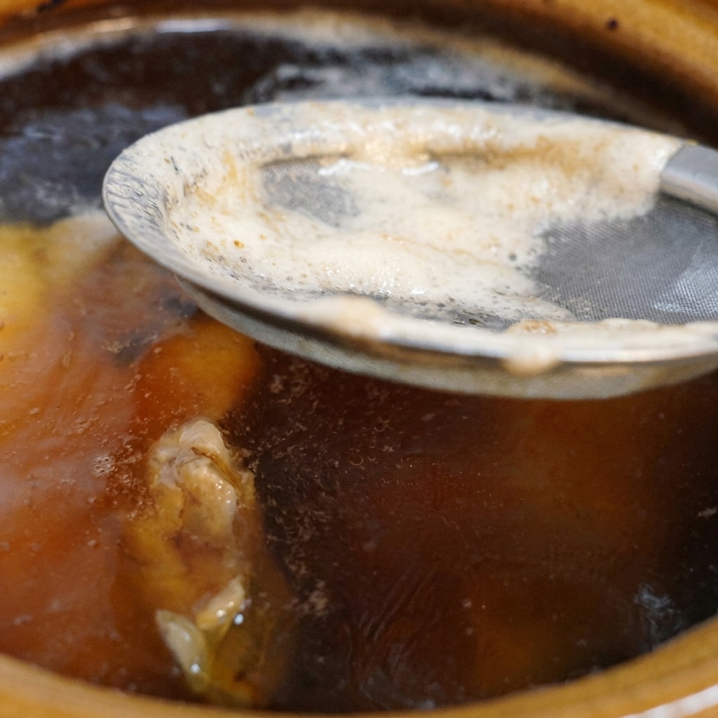 Une passoire métallique à mailles fines retirant de lécume blanche à la surface dun bouillon de poulet foncé qui bouillonne dans une marmite en céramique.
