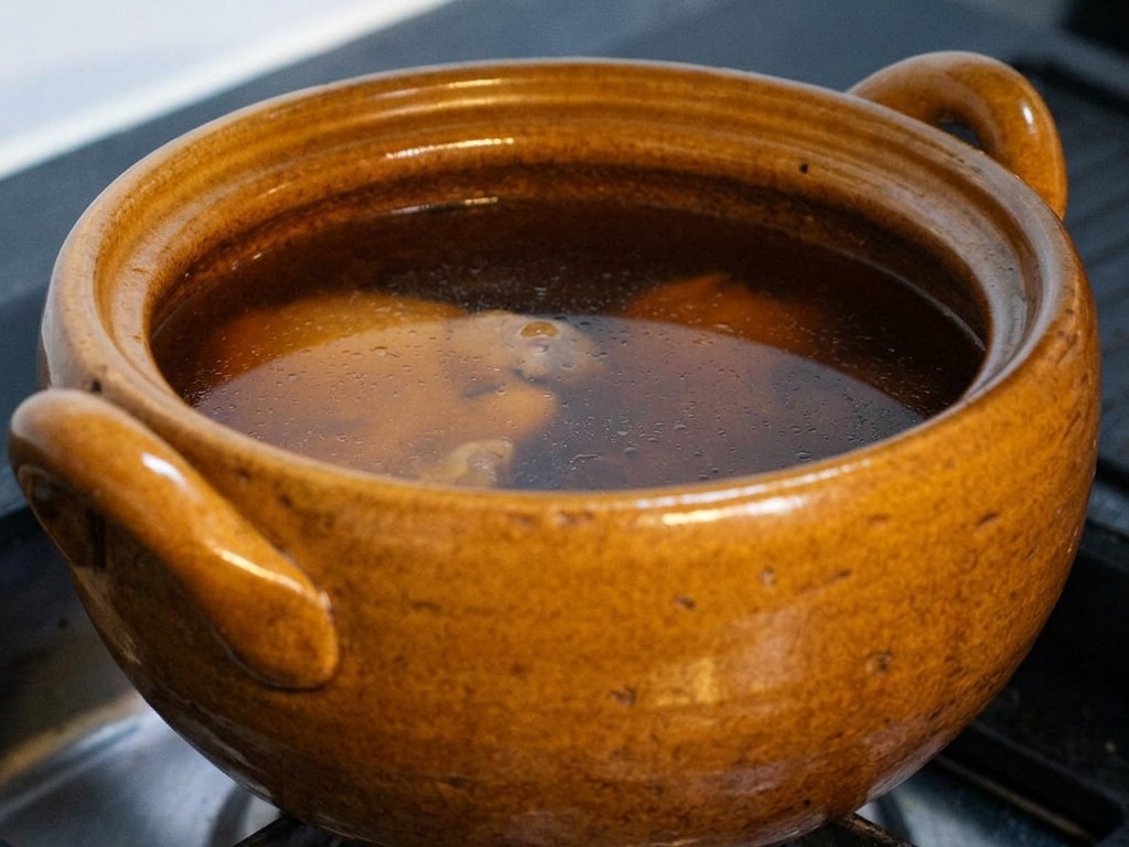 Une marmite en céramique brune sur une gazinière remplie dun bouillon foncé à base de sauce soja frémissant doucement, le couvercle étant partiellement soulevé pour laisser entrevoir le poulet et le kombu.