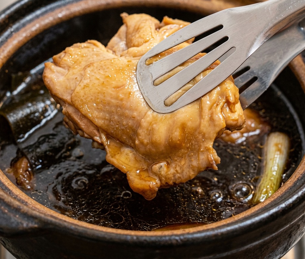 Des pinces métalliques sortant un morceau de poulet cuit et bien coloré dune marmite de bouillon de volaille foncé.