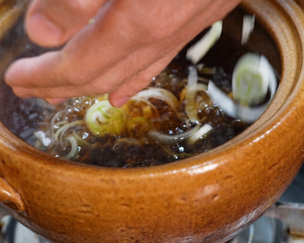 Des mains déposant une poignée doignons verts émincés dans une marmite de bouillon de poulet sombre frémissant.