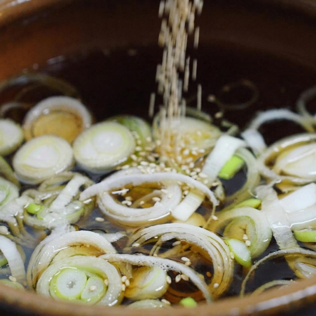 Des graines de sésame blanc tombant dans un bouillon sombre frémissant rempli doignons verts émincés dans une marmite en céramique.