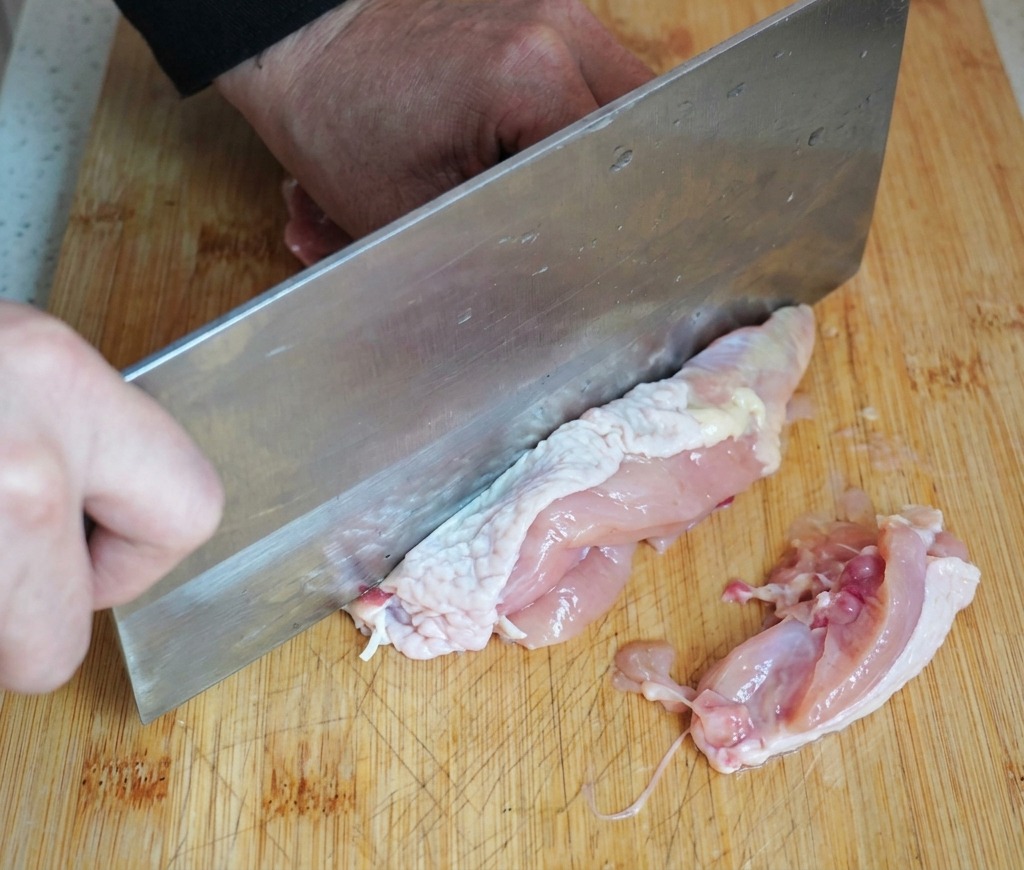 Une personne utilisant un grand couperet en métal pour désosser un haut de cuisse de poulet cru sur une planche à découper en bois.