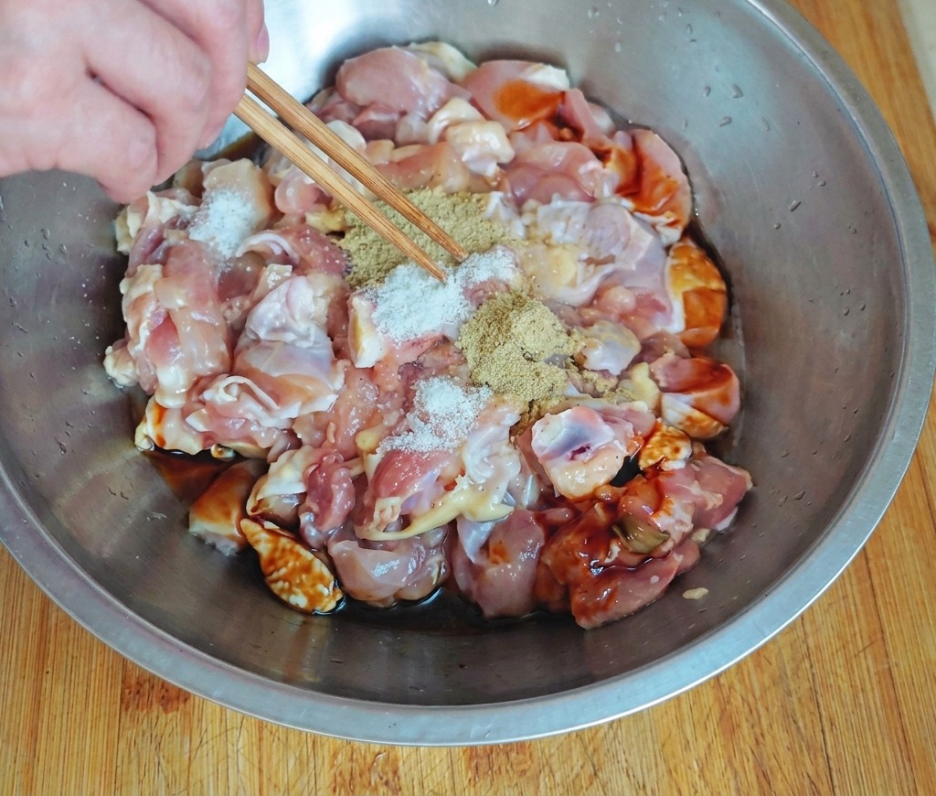 Une main utilisant des baguettes pour saupoudrer des épices blanches sur des morceaux de poulet cru dans un bol en acier inoxydable contenant des sauces foncées.