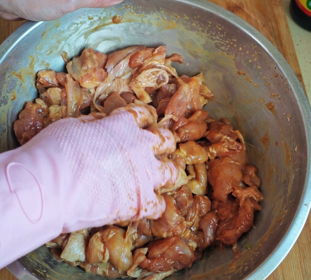 Une main portant un gant rose mélangeant soigneusement des morceaux de poulet marinés avec de la fécule et de lhuile dans un grand bol en acier inoxydable.