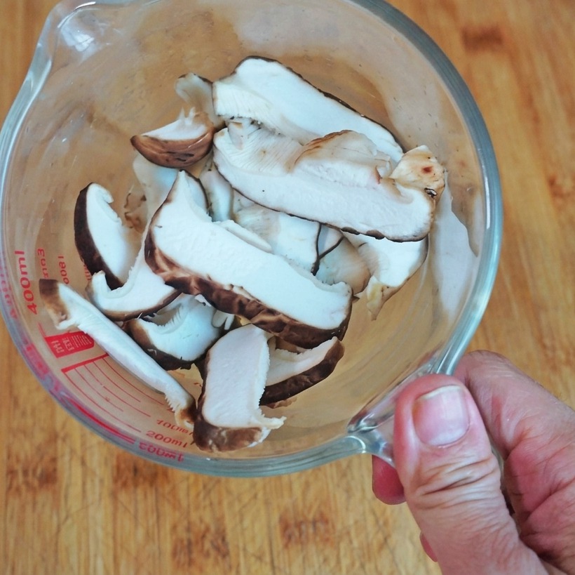 Une vue de dessus de champignons shiitakés frais émincés dans un verre doseur transparent posé sur une planche en bois.
