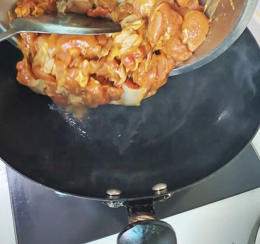 Des morceaux de poulet mariné versés dun bol en métal dans un wok noir chaud et fumant.