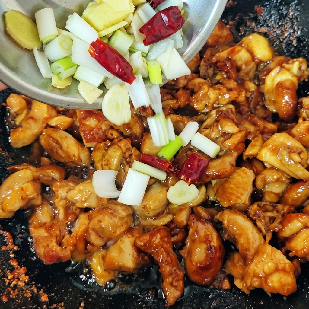 Un mélange de ciboules émincées, de tranches de gingembre et de piments rouges séchés versé dun petit bol dans un wok rempli de morceaux de poulet saisis.