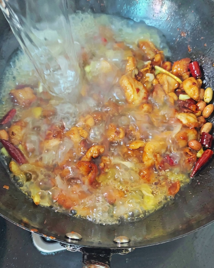 Un filet deau bouillante versé dans un wok noir contenant du poulet saisi et des piments rouges, créant un bouillon bouillonnant.