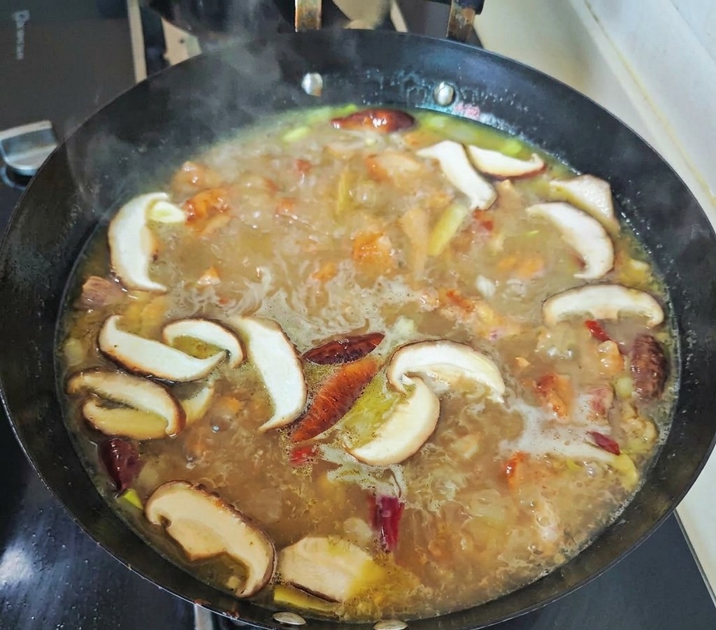 Des tranches de shiitakés et des piments rouges mijotant dans un bouillon de poulet doré et bouillonnant à lintérieur dun wok noir.
