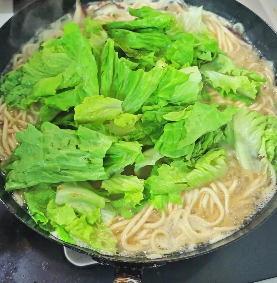 Une grande poignée de légumes verts à feuilles frais et de pousses dail déposée sur les nouilles dans un wok de soupe en ébullition.