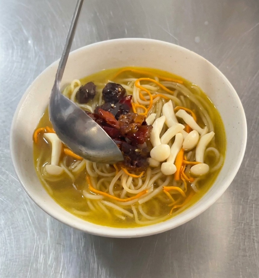Une louche en métal déposant des morceaux de poulet sauté mariné en rouge et des piments séchés sur un bol de nouilles au bouillon doré.