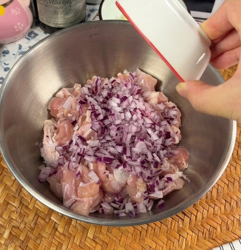 Une main versant un petit bol doignons rouges émincés et dail haché sur des morceaux de poulet cru dans un saladier en métal.