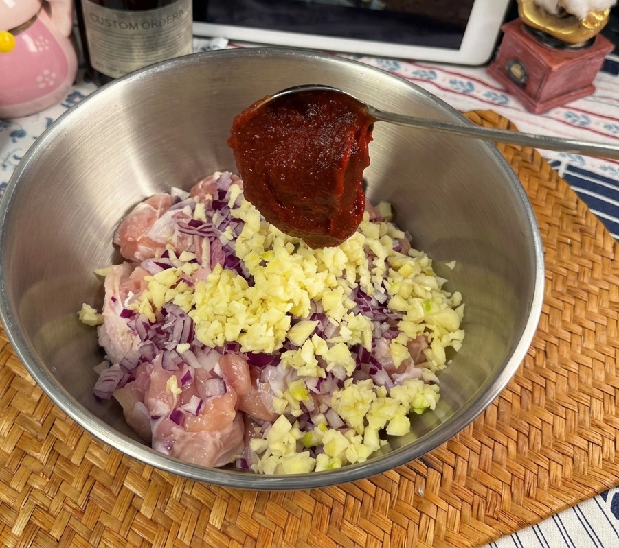 Une cuillère en métal contenant une généreuse portion de pâte de Gochujang rouge vif au-dessus dun bol de poulet cru, dail et doignons.