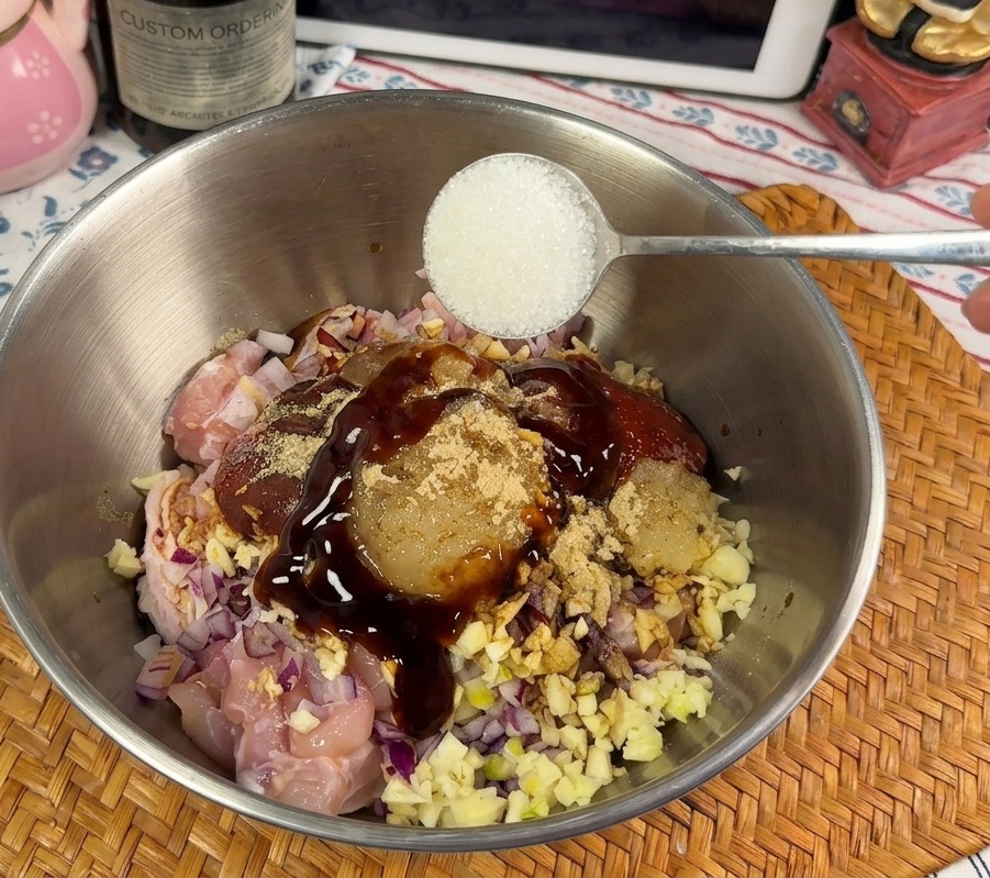 Une cuillérée de sucre blanc en poudre ajoutée à un bol de poulet marinant dans une sauce rouge épaisse.