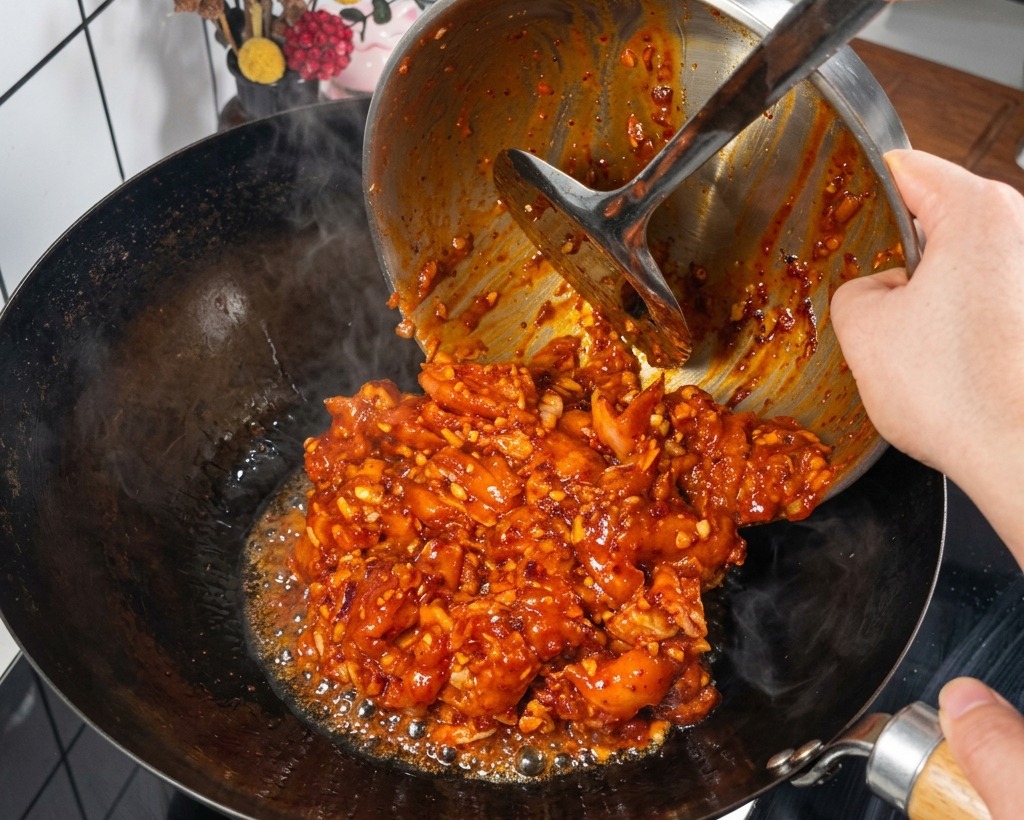 Des morceaux de poulet mariné dans une sauce rouge vif versés depuis un bol en métal dans un wok noir chaud et fumant.