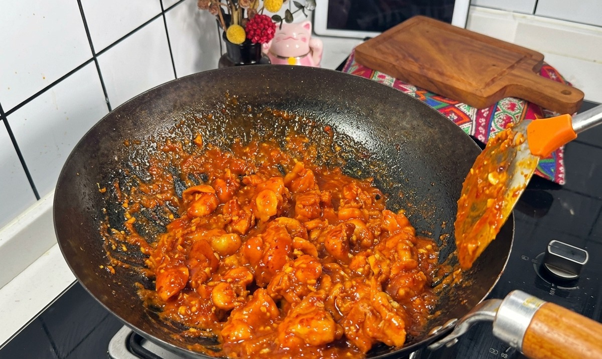 Des morceaux de poulet enrobés dune sauce rouge épaisse et bouillonnante sautés dans un wok noir à laide dune spatule en métal.