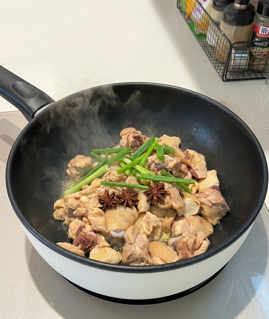 Des étoiles de badiane entières et des tiges doignons verts déposées sur des morceaux de poulet dans une poêle fumante.