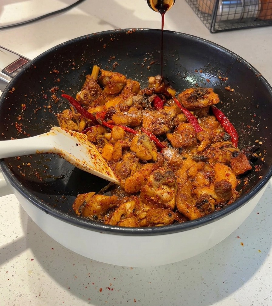 De la sauce soja foncée versée depuis une bouteille dans une poêle contenant des morceaux de poulet épicés.