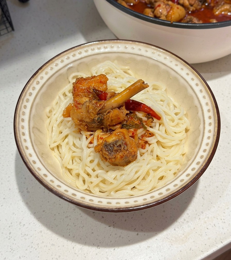 Un bol en céramique de nouilles blanches surmonté de poulet épicé et de sauce au piment rouge, avec la poêle visible en arrière-plan.