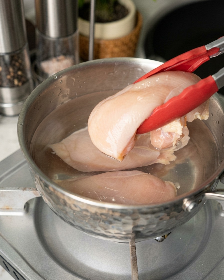Une personne utilisant des pinces aux embouts rouges pour plonger des blancs de poulet crus et roses dans une casserole deau en acier inoxydable.