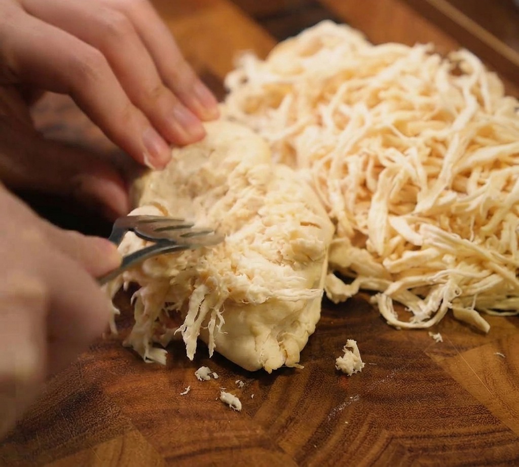 Une paire de fourchettes métalliques utilisée pour effilocher un blanc de poulet cuit en fines lamelles sur une planche à découper en bois foncé.
