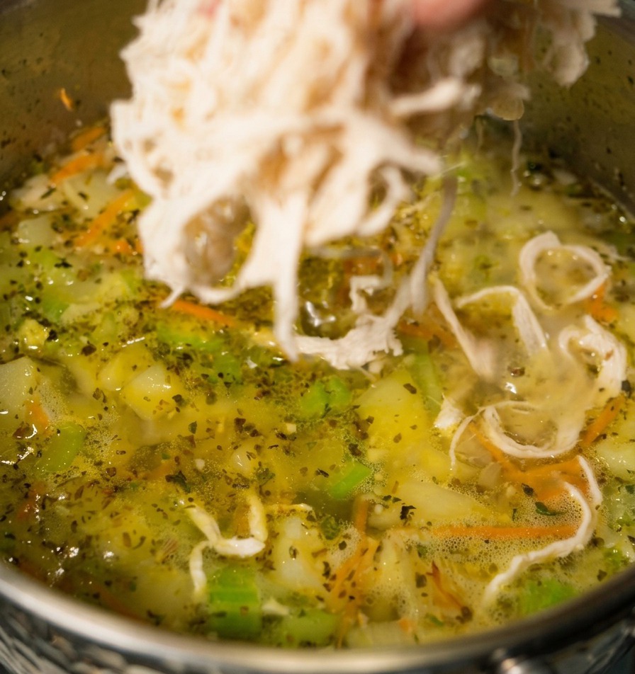 Une main déposant du poulet blanc effiloché dans une marmite de soupe de légumes frémissante où lon distingue des carottes et du céleri.