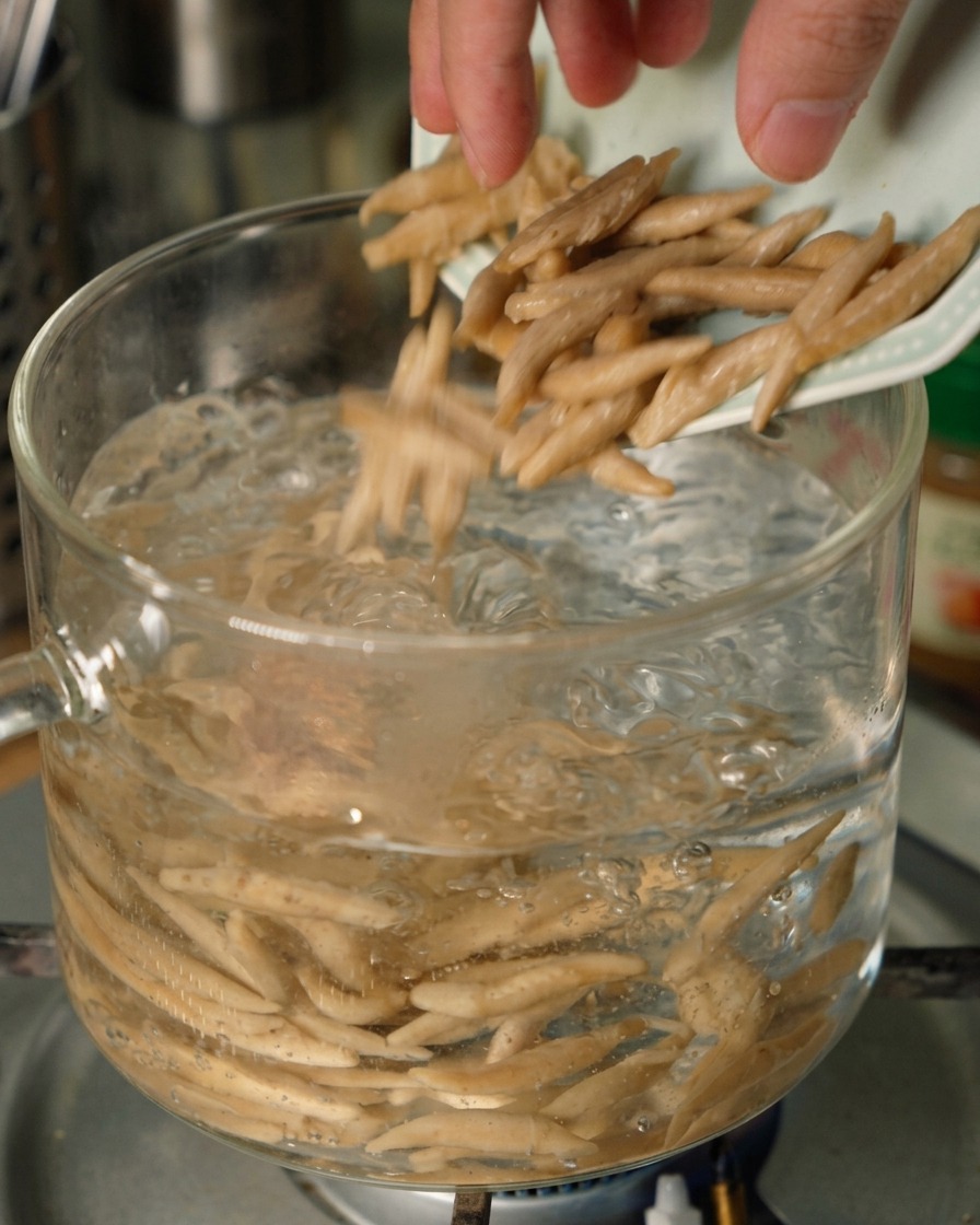 Des nouilles davoine effilées glissant dune petite assiette dans une casserole en verre transparent remplie deau bouillante à gros bouillons.