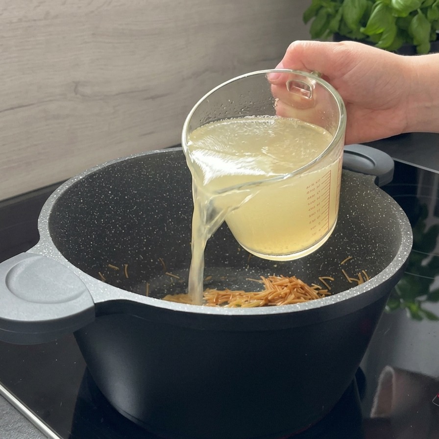 Un bouillon de poulet clair versé depuis un pichet en verre dans une marmite noire contenant les pâtes torréfiées.