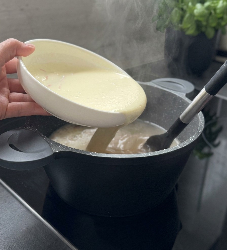 Une liaison blanche et crémeuse versée dun bol blanc dans une marmite de soupe fumante tout en étant remuée avec une spatule noire.