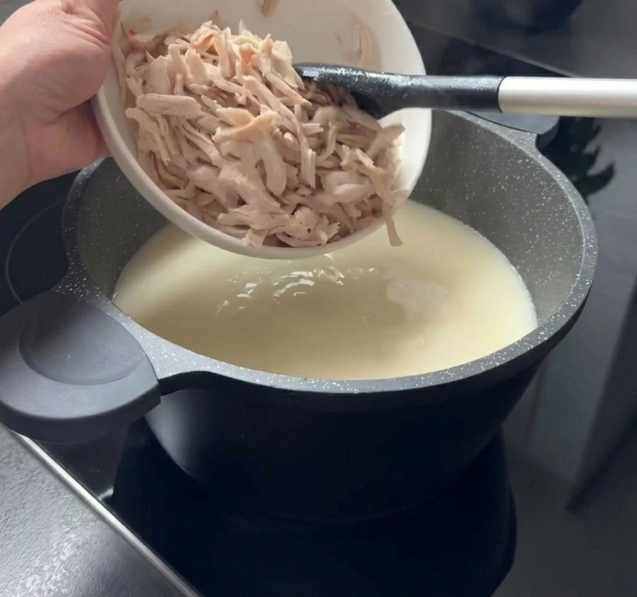 Des morceaux de poulet cuit et effiloché ajoutés depuis un bol blanc dans une marmite de soupe blanche crémeuse.