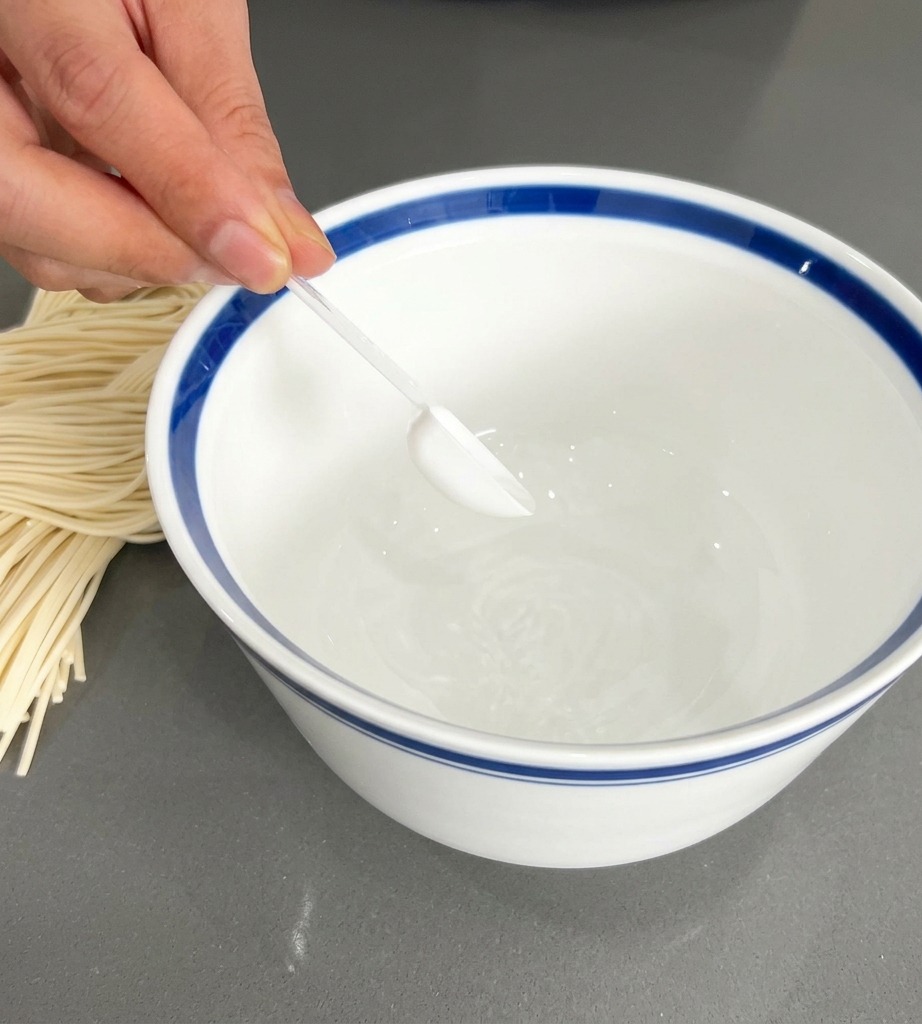 Une main tenant une petite cuillère transparente, déposant du bouillon en poudre blanc dans un bol blanc au bord bleu distinctif.