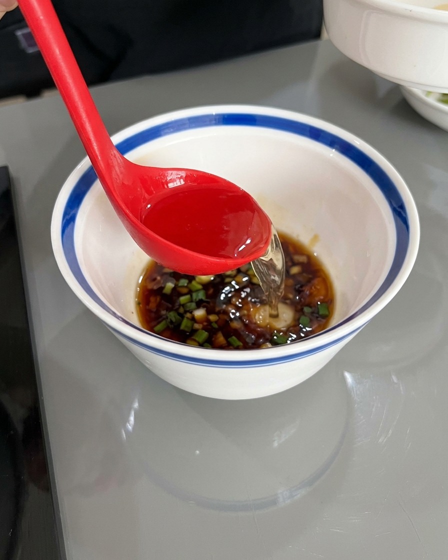 Une louche rouge versant de leau chaude et claire dans un bol blanc contenant la sauce soja foncée, lhuile et les aromates émincés.