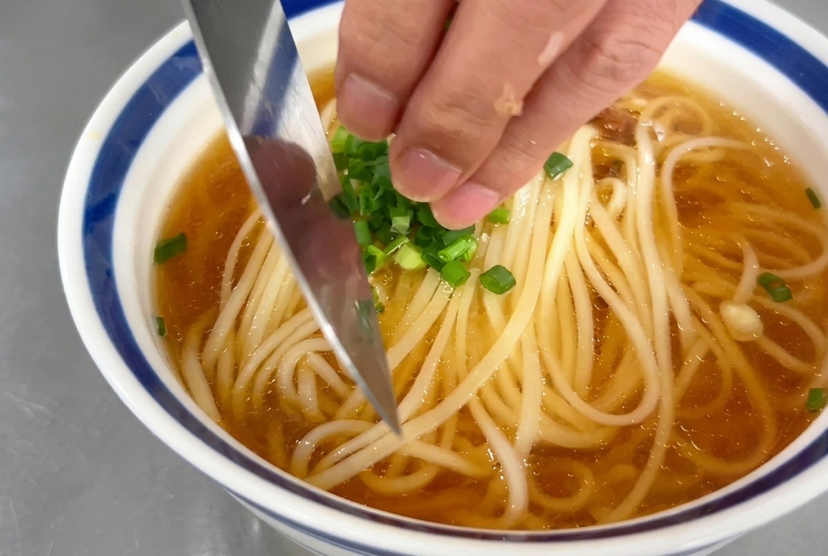 Une main parsemant de fines rondelles doignons verts sur un bol de soupe de nouilles.