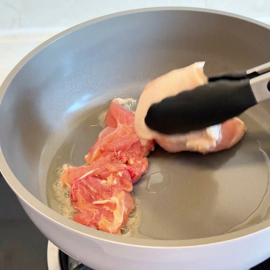Une pince déposant un haut de cuisse de poulet cru côté peau dans une poêle antiadhésive grise avec de lhuile chaude.