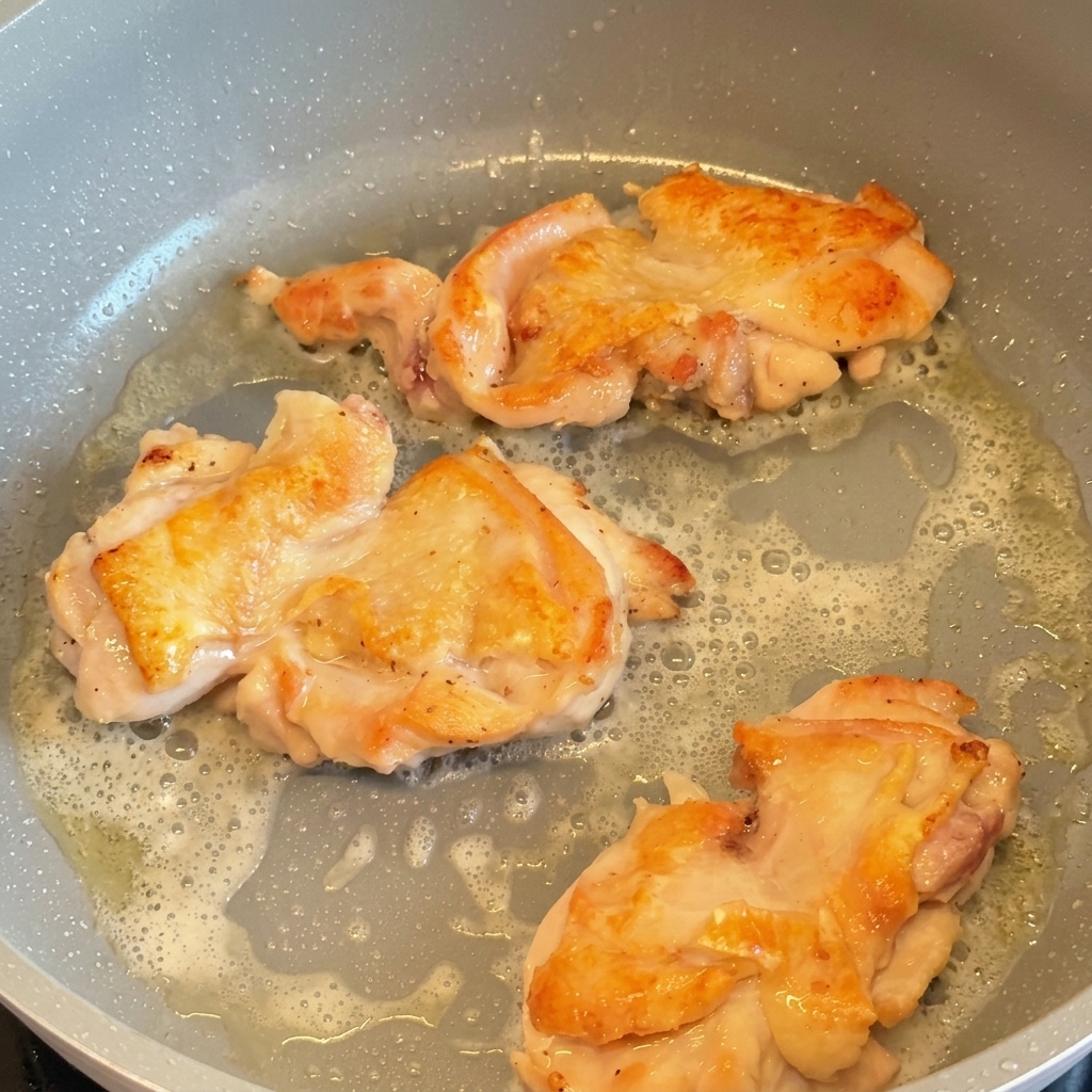Trois hauts de cuisse de poulet dorant dans une poêle, retournés pour révéler leur peau croustillante et dorée.