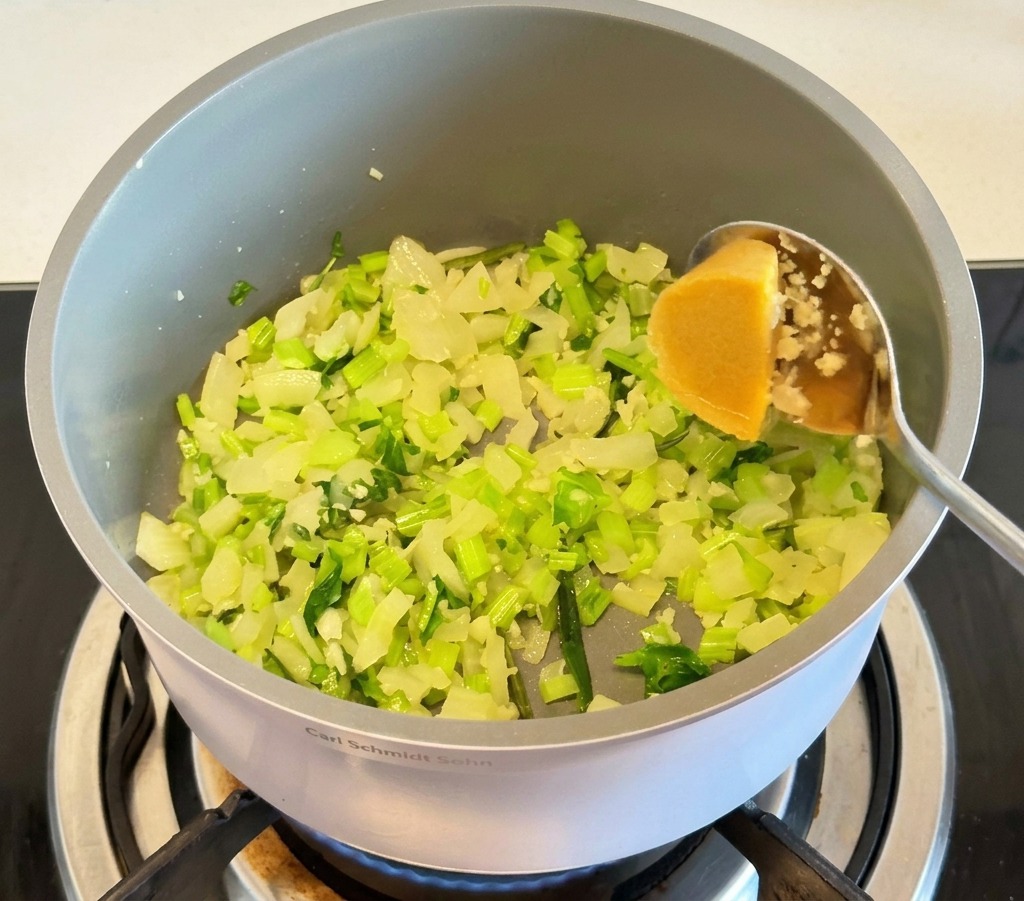 Une cuillère en métal ajoutant un demi-cube de bouillon de poulet concentré dans une marmite grise avec les oignons et le céleri.