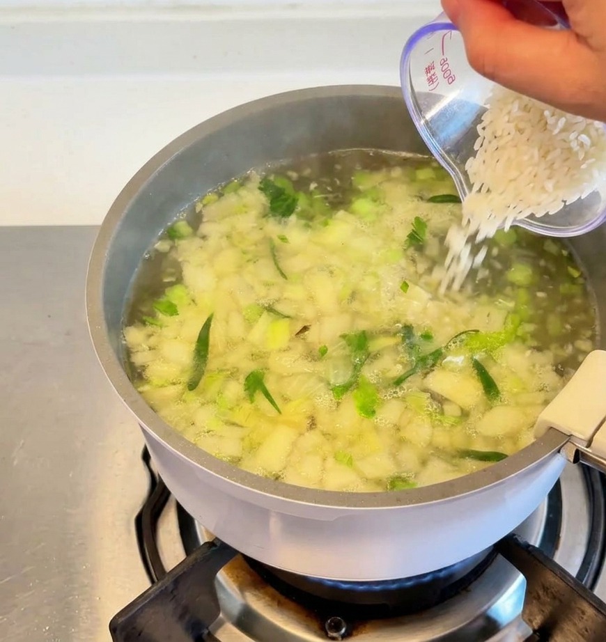 Du riz blanc versé dun verre doseur transparent dans une marmite de soupe frémissante parsemée de morceaux de légumes.