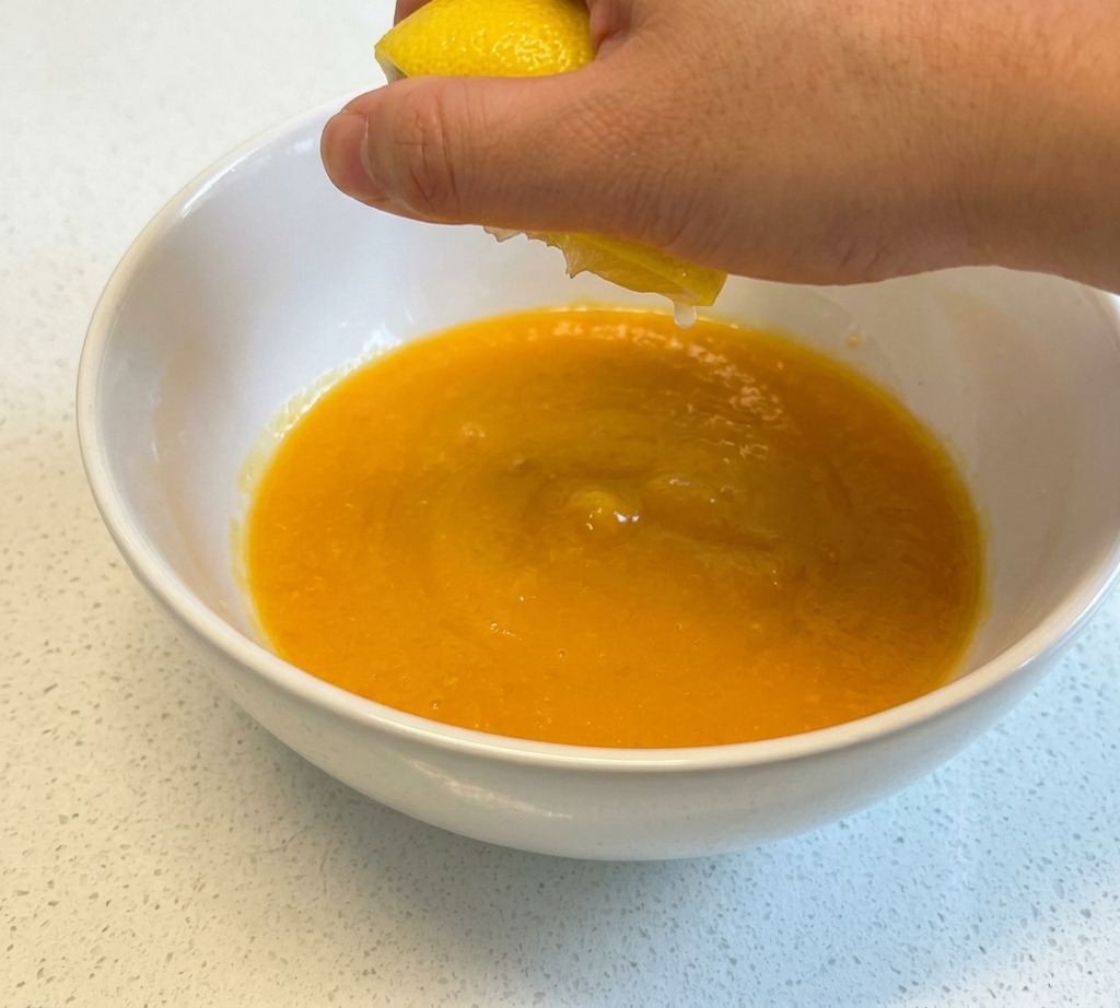 Une main pressant le jus dun demi-citron jaune au-dessus dun bol en céramique blanche contenant des œufs battus.