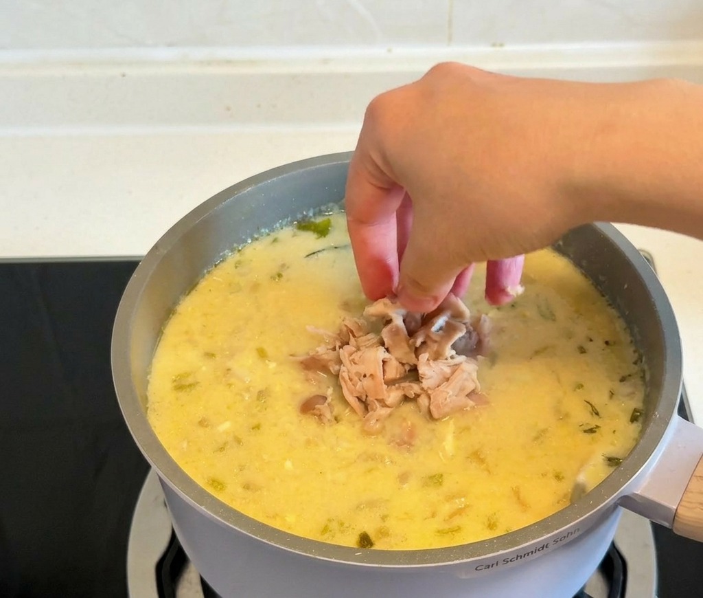 Des mains déposant des morceaux de poulet cuit effiloché dans une marmite de soupe jaune crémeuse et liée.