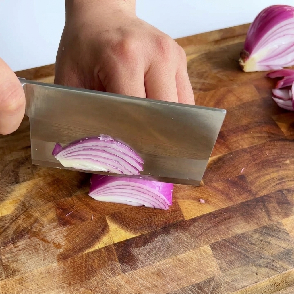 Un grand couperet tranchant un oignon rouge pour créer de fines lamelles violettes sur une planche.