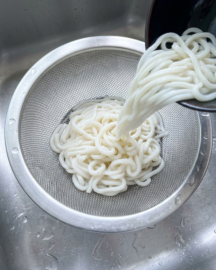 Nouilles udon blanches versées dans une passerelle en inox sous un jet deau froide dans un évier.