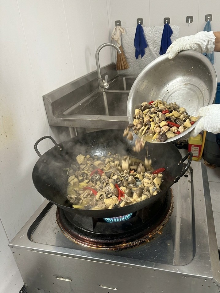 Une personne versant de leau bouillante limpide depuis un récipient dans un wok fumant contenant le poulet saisi et les épices.