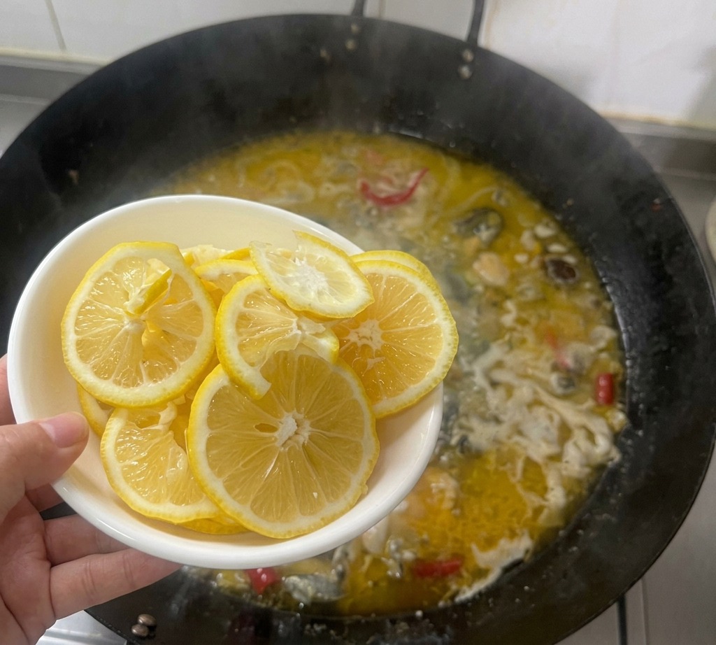 Une main tenant un bol blanc rempli de fines tranches de citron au-dessus dun wok où mijote un bouillon de poulet doré.