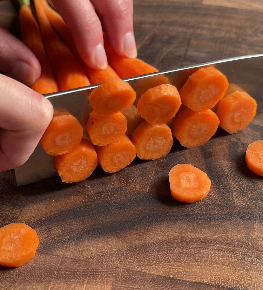 Gros plan sur des mains coupant plusieurs carottes en fines rondelles avec un couteau sur une planche en bois sombre.