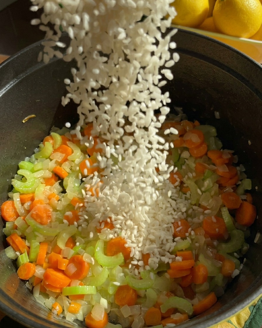 Un filet de riz jasmin blanc versé dans une marmite sombre remplie de carottes, de céleri et doignons revenus.