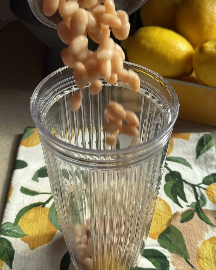 Haricots blancs cuits tombant dans le bol dun blender transparent posé sur un torchon aux motifs de citrons.