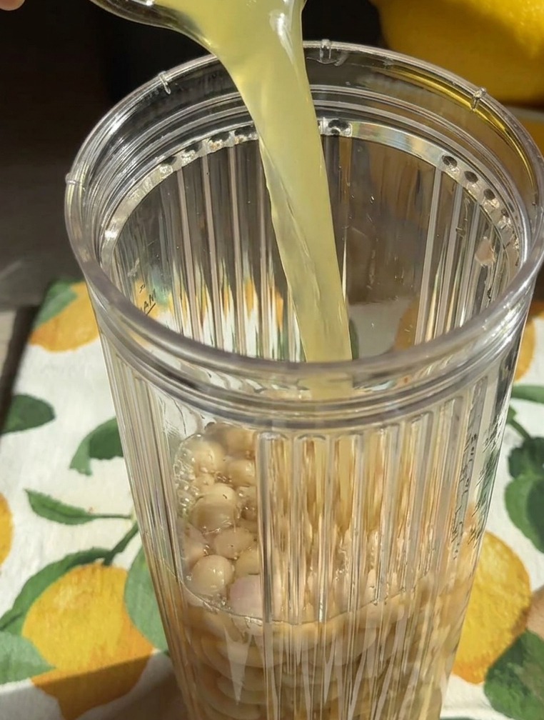 Un liquide jaunâtre versé depuis un verre dans le bol dun blender rempli de haricots blancs entiers.